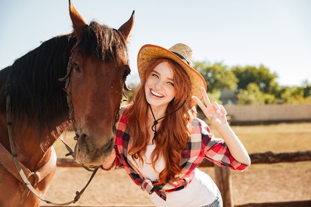 Happy lovely young woman cowgirl standing with horse and showing peace signの写真素材