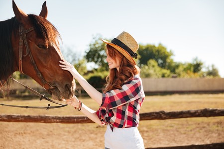 Smiling redhead young woman cowgirl taking care of her horse on farmの写真素材