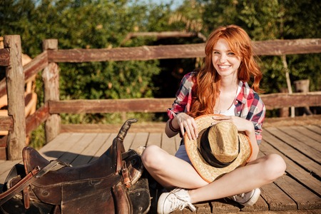 Happy beautiful young woman cowgirl with hat sitting on ranch