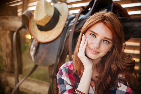 Close up portrait of a happy beautiful redhead cowgirl resting at the ranch fenceの写真素材