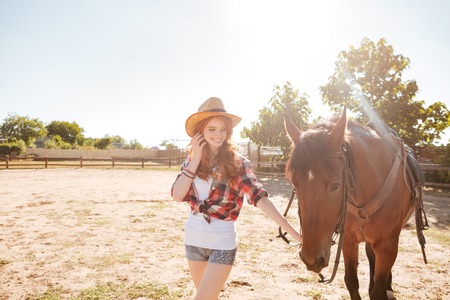 Cheerful beautiful young woman cowgirl walking with her horse on ranchの写真素材