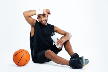 Tired young man basketball player sitting and drinking water over white backgroundの写真素材