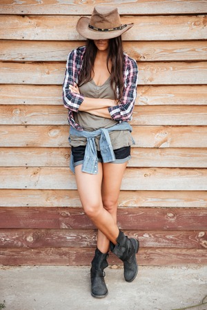 Beautiful young woman cowgirl covered face by hat standing with arms crossedの写真素材