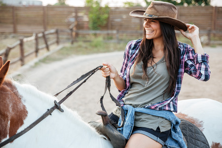 Cheerful attractive young woman cowgirl smiling and riding horse in villageの写真素材