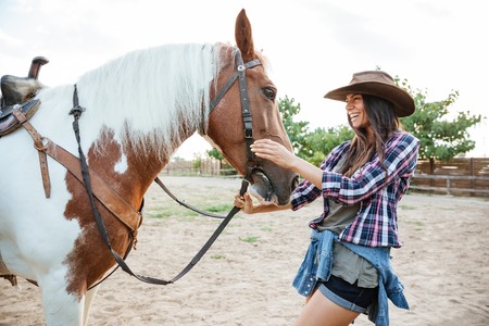 Cheerful charming young woman cowgirl with horse standing and laughingの写真素材
