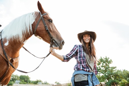 Happy smiling young woman in cowboy hat taking her horse for a walk at the farmの写真素材
