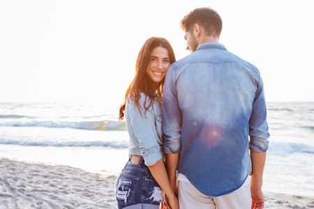 Beautiful young woman holding her boyfriend hand who standing backwards at the beachの写真素材