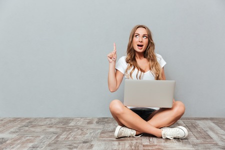 Portrait of a smiling woman sitting on the floor with laptop and pointing finger up isolated on gray backgroundの写真素材