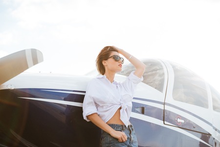 Beautiful young woman in sunglasses standing outdoors near small private aircraft