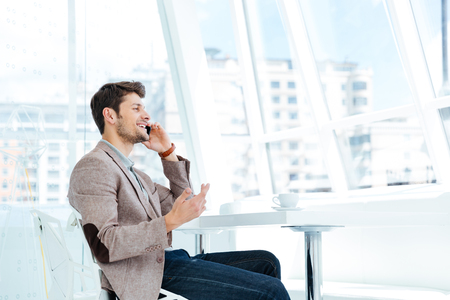 Happy smiling casual businessman talking on mobile phone while sitting at the table indoorsの写真素材