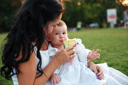 Cute small girl sitting and drinking juice with her mom outdoorsの写真素材