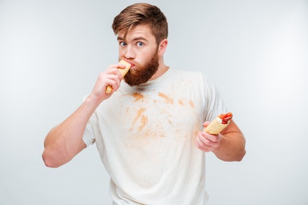 Hungry bearded man in dirty shirt biting two hotdogs isolated on white backgroundの写真素材