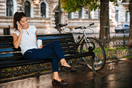 Beautiful casual girl talking on the mobile phone while sitting on the bench with cup of coffeeの写真素材