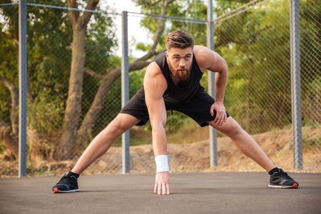 Confident young bearded male athlete stretching and warming up before jogging outdoors