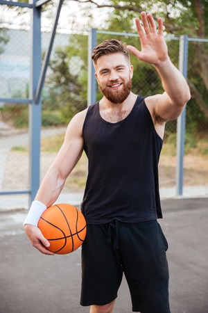 Young smiling bearded basketball player greeting somebody with hand waving outdoorsの写真素材