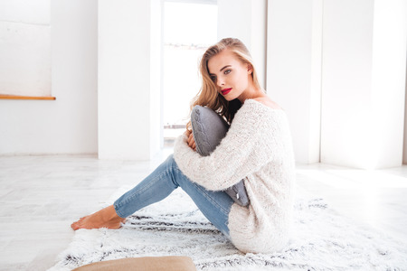 Beautiful charming woman wearing sweater and red lipstick sitting on the carpet at homeの写真素材