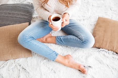 Cropped image of a woman in sweater and jeans holding tea cup while sitting with legs crossed indoorsの写真素材