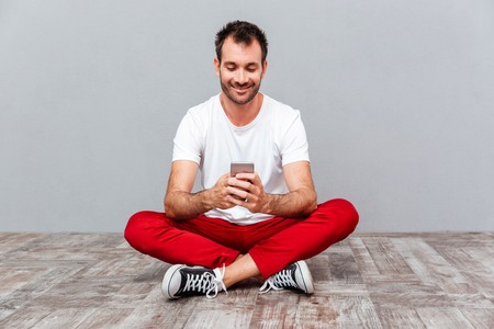 Happy casual man sitting on the floor and using smarpthone over gray background