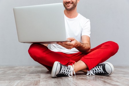 Cropped image of a happy smiling casual man sitting on the floor with laptop over gray backgroundの写真素材