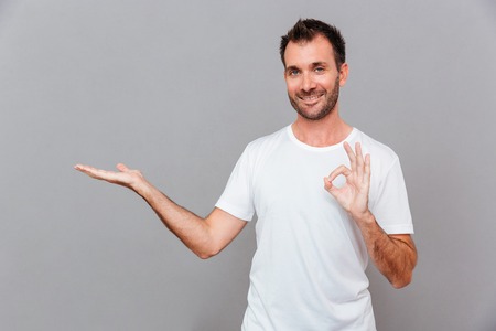 Attractive happy smiling casual young man in white t-shirt holding copyspace on palm and showing okay gesture over gray backgroundの写真素材