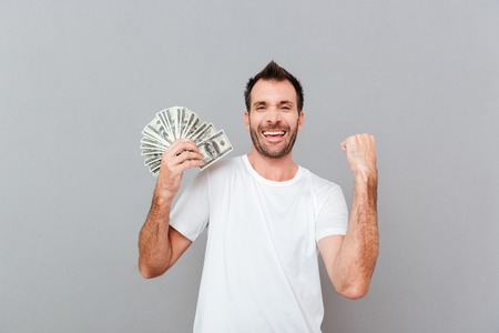 Cheerful excited young man holding dollars and celebrating success over gray backgroundの写真素材