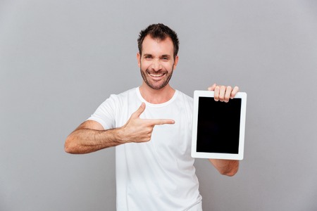 Portrait of a handsome man pointing finger on blank tablet computer screen isolated on a gray backgroundの写真素材