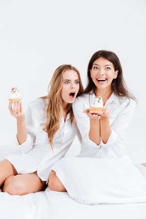 Two happy smiling women in white shirts eating sweet cupcakes in bedの写真素材