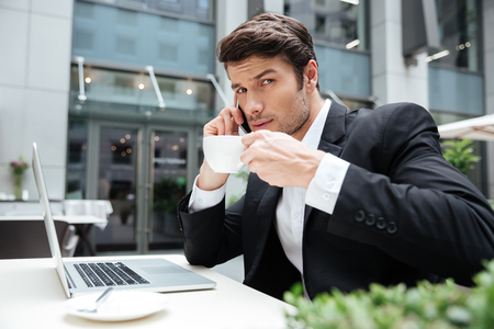 Businessman with laptop talking on mobile phone and drinking coffee in outdoor cafeの写真素材