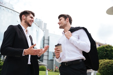 Two cheerful young businessmen talking and drinking coffee outdoors