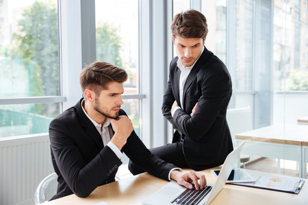 Two serious young businessmen working and using laptop in office togetherの写真素材