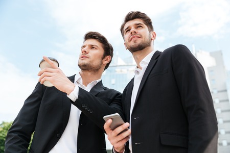 Two confident young businessmen with take away coffee and cell phone outdoorsの写真素材