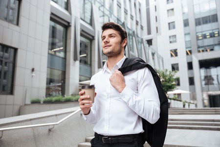 Handsome young businessman with jacket and cup of coffee walking near business centerの写真素材