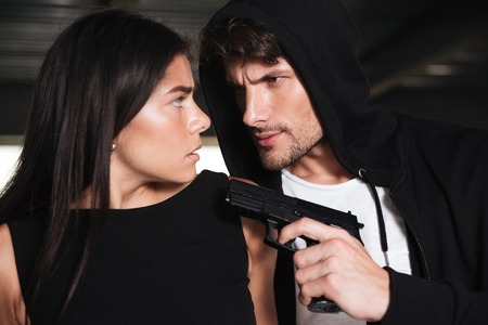 Closeup of scared young woman threatened with gun by criminal man in hoodieの写真素材