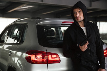 Criminal man in black hoodie with gun standing near car on parking