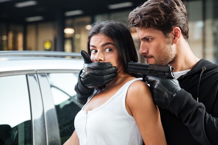 Dangerous young man robber in gloves covered mouth of woman and threatening with gun outdoorsの写真素材