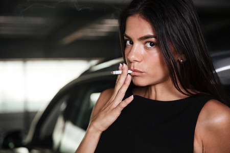 Beautiful young woman smoking cigarette on car parkingの写真素材