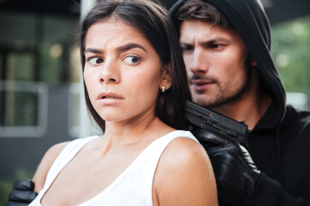 Man robber in hoodie threatening with gun to terrified young woman outdoorsの写真素材