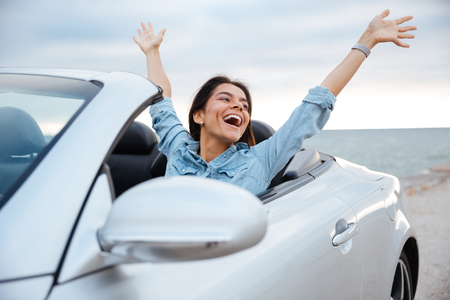 Young attractive brunette woman sitting with her hands up in cabrioletの写真素材