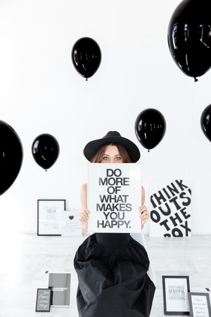 Cute amusing young woman in hat sitting and hiding behind white board over black balloons background