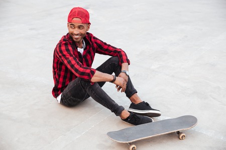 Photo of cheerful dark skinned boy wearing cap sitting near his skateboard. Against nature background.の写真素材