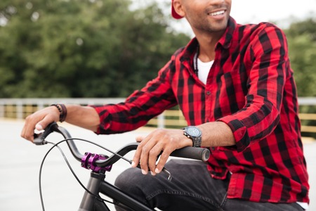 Cropped picture of happy dark skinned guy wearing cap sitting on his bicycle. Against nature background.の写真素材