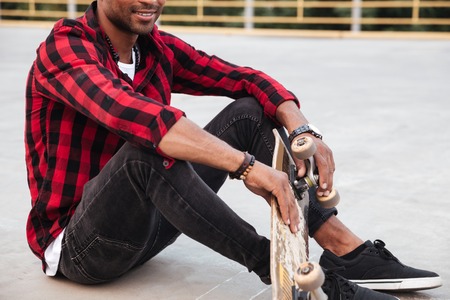 Cropped photo of dark skinned young man sitting near his skateboard. Against nature background.の写真素材