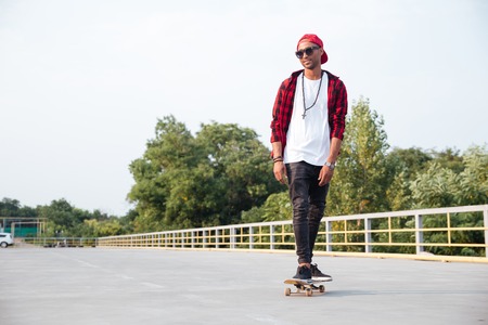 Picture of young dark skinned man wearing sunglasses and cap skateboarding. Against the nature background.の写真素材