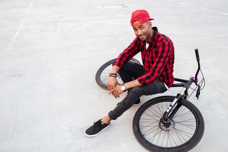 Photo of cheerful dark skinned guy wearing cap sitting on his bicycle. Against nature background. Looking at the camera.の写真素材