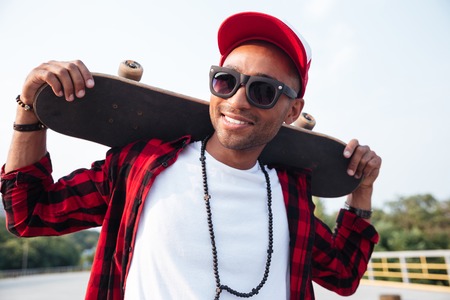 Photo of young dark skinned man wearing sunglasses holding skateboard. Against the nature background.の写真素材