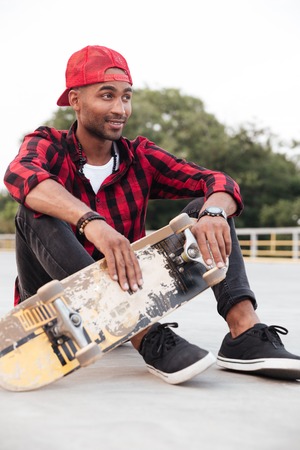 Picture of happy dark skinned young man wearing cap sitting and holding his skateboard. Against nature background.の写真素材