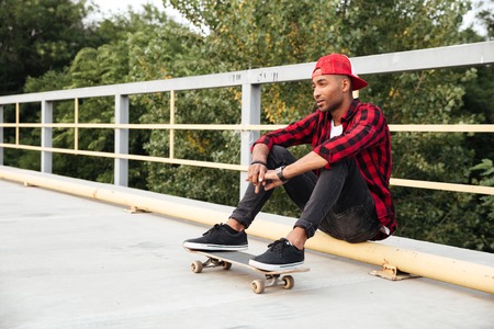 Picture of young dark skinned man wearing sunglasses and cap sitting with his skateboard. Against the nature background.の写真素材