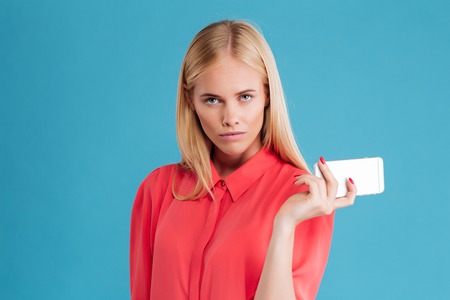 Serious pretty blonde girl holding mobile phone and looking at camera over blue backgroundの写真素材