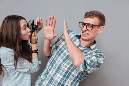 Irritated young man in eyeglasses covers with hands from annoying woman photograher isolated on the gray backgroundの写真素材