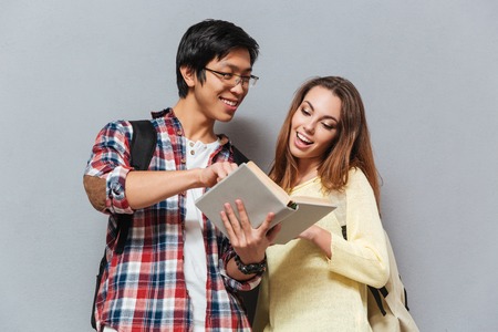 Portrait of a smiling multicultural couple with backpacks reading book isolated on the gray backgroundの写真素材